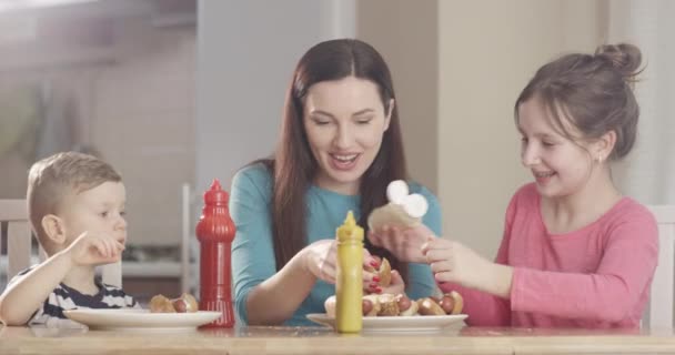 お母さんと一緒にホットドッグを作る子供たち一緒に笑顔で楽しい時間を過ごすと台所のテーブルに座って食べ物を楽しむ — ストック動画