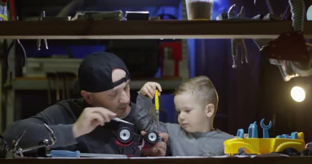 父と息子がガレージでドライバーとおもちゃのトラクターを一緒に固定し 小さな男の子は赤いカメラで撮影されたツールで動作するように学習しています — ストック動画