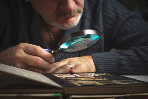 Man considers album of postage stamps trough the magnifying glass horizontal — Stock Photo, Image