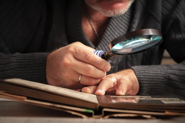 Man considers album of stamps trough the magnifying glass horizontal — Stock Photo, Image