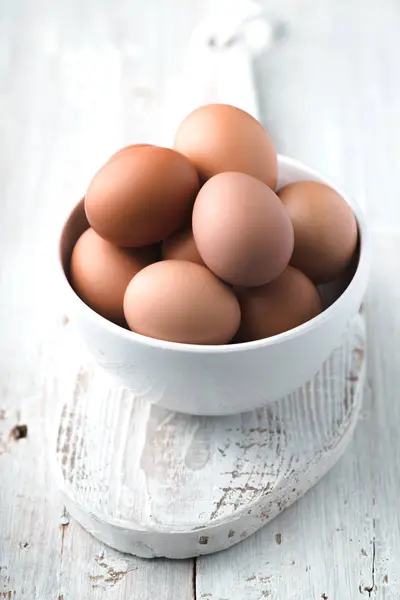 Куриные яйца в керамической миске на белом деревянном столе вертикальный — стоковое фото