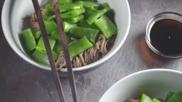 Гречневая лапша с зеленой фасолью в керамических мисках и соевым соусом на сером камне — стоковое видео