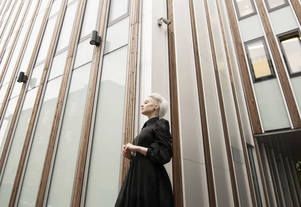 Wide angle view, woman in long black dress with short white haircut, building