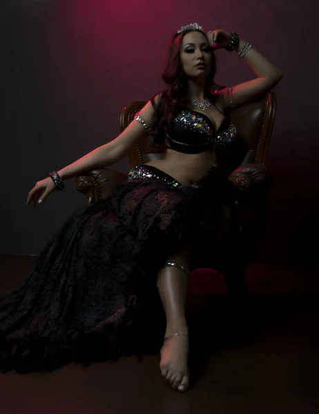 クリスタルと贅沢な茶色の革の椅子、壁にピンクの光の羽で黒のステージ衣装で完璧なボディのセクシーな女性, — ストック写真