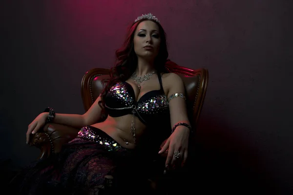 Sexy bellydancer woman in black stage costume with crystals and feathers on luxury brown leather chair like a boss — Stock Photo, Image