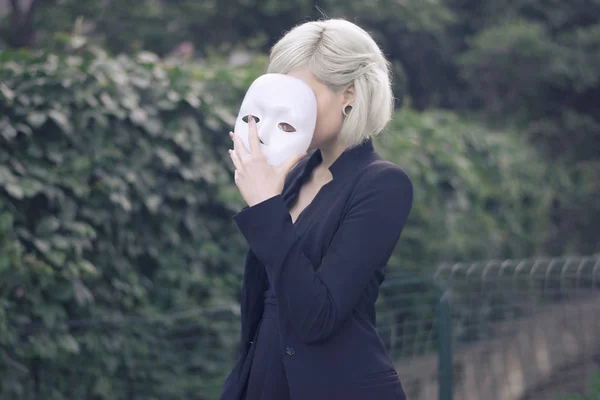 Young blond girl taking off a mask. Pretending to be someone else concept. outdoors. — Stock Photo, Image