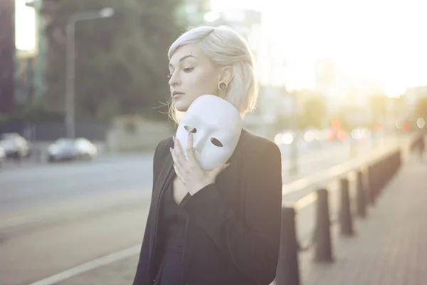 Young blond woman taking off a mask. Pretending to be someone else concept. outdoors on sunset. — Stock Photo, Image