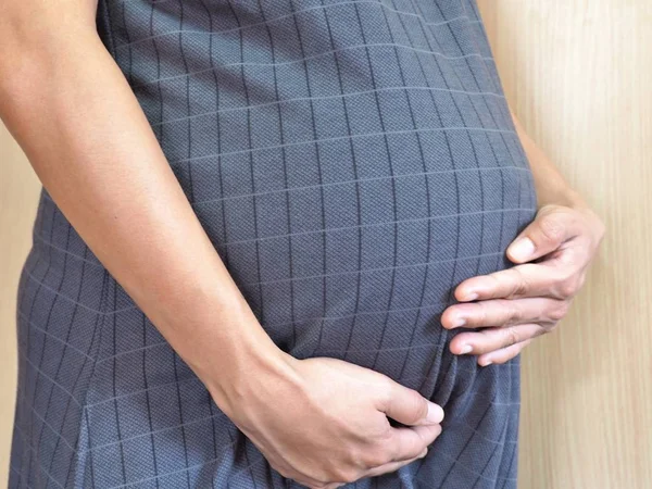 Close-up of Asian pregnant woman in maternity clothes holds hands on belly on wooden background. Image of pregnancy and maternity concept.