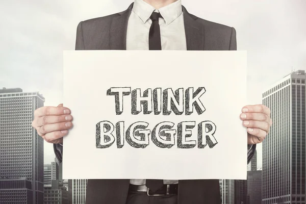 Businessman Holding Placard With Think Bigger Text Against Buildings — Stock Photo, Image