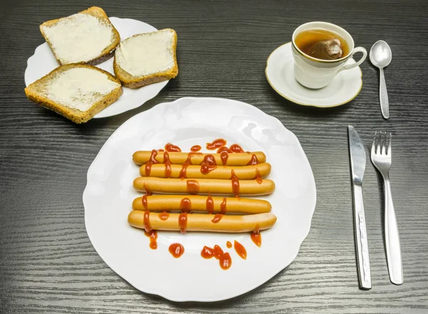 Ein Teller Kochwürste mit Ketchup, Sandwiches mit dunkelbraunem Brot mit Butter und eine Tasse aufgebrühten Tee aus der Tüte. — Stockfoto