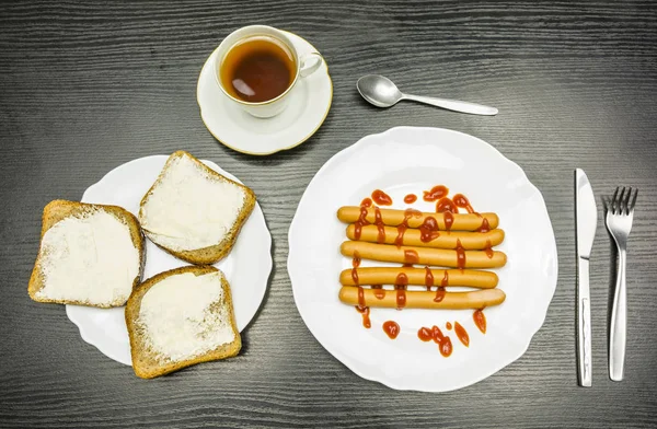 Frühstück - gekochte Würstchen mit Ketchup, Scheiben mit Butter und eine Tasse Tee. Blick von oben. — Stockfoto