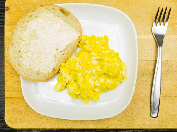 Teller mit Rührei und einer Scheibe Brot mit Butter auf einem Holzbrett. Blick von oben. — Stockfoto