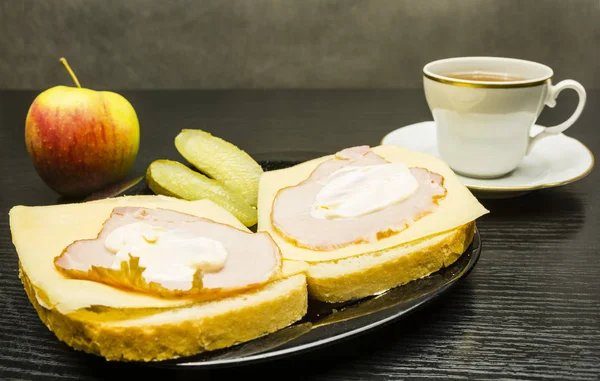 Een plaat met broodjes met ham, kaas en mayonaise en komkommer. Een appel en een kopje thee. — Stockfoto