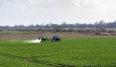 Nieciecza, Polonya - 28 Mart 2020: Mavi Hollanda traktörü ve yeşil püskürtücü Krukowiak bahar zamanı tecavüz tohumları püskürtülürken.