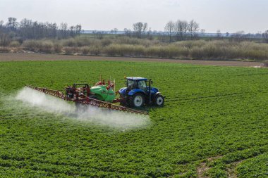 Nieciecza, Polonya - 28 Mart 2020: Tarım. Mavi Hollanda T6 traktörü yeşil ve kırmızı püskürtücü Krukowiak Goliat bahar kolza tohumu püskürtürken.