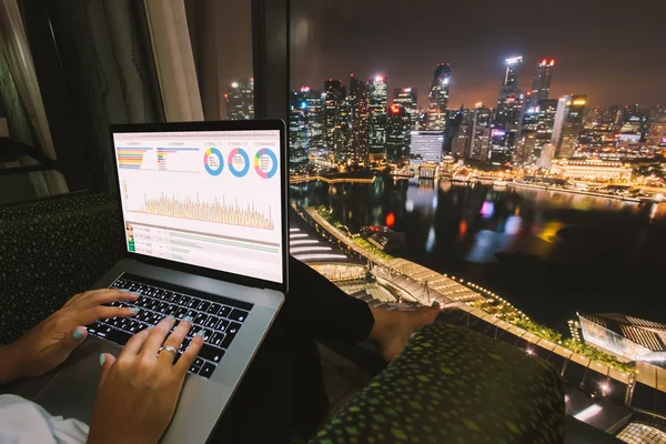 Woman freelancer is working using laptop computer in home office at night. Aerial city skyline view from window. Freelance lifestyle