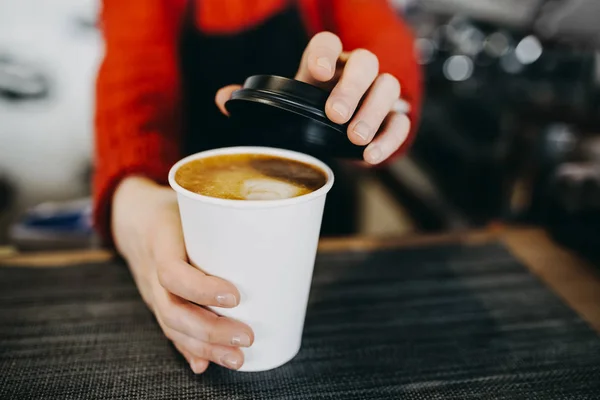 Barista em avental está segurando em mãos cappuccino quente em copo de papel branco takeaway. Café levar na loja de café — Fotografia de Stock