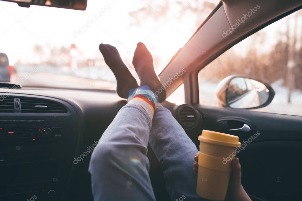 Woman is holding cup of coffee inside of car. Travel lifestyle. Legs on dashboard