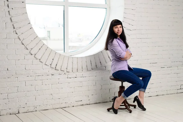 Beautiful girl sits in an office on a chair near a large round window — Stock Photo, Image