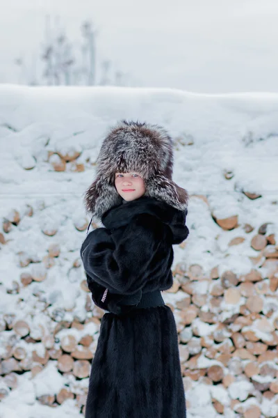 Ragazza in un cappello di pelliccia con paraorecchie e una pelliccia in piedi presso la catasta di legno — Foto Stock