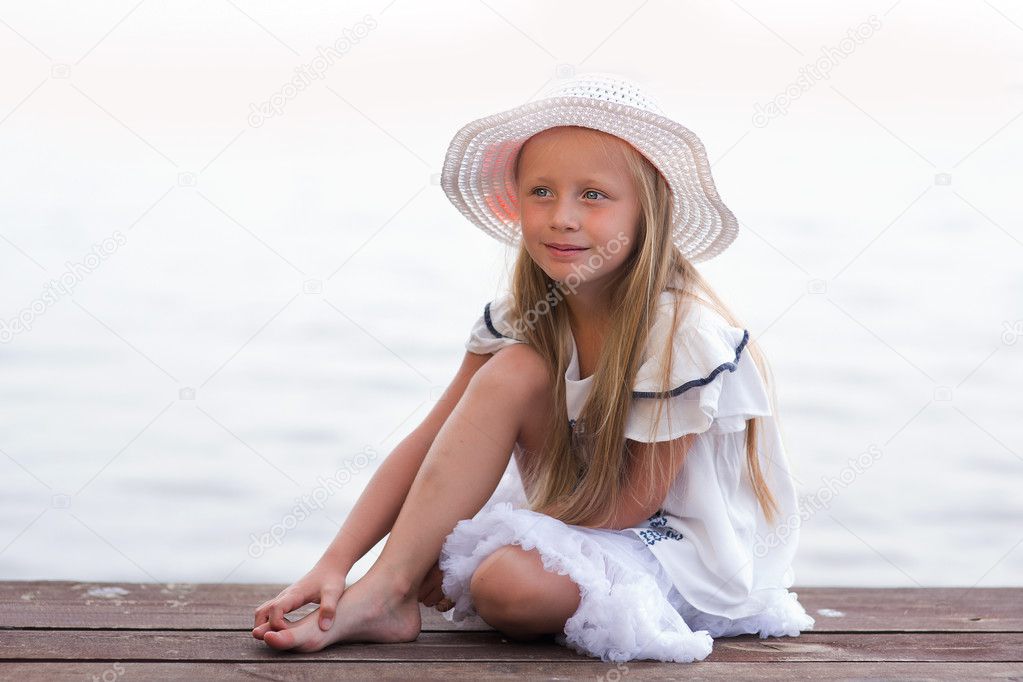 beautiful girl in a white hat and white dress sitting on the pier at the lake