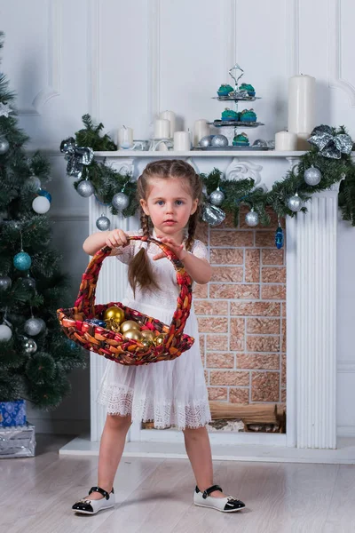 Маленькая девочка с двумя косичками, стоящая рядом с елкой. Она держит корзину с игрушками — стоковое фото