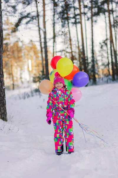 Mała dziewczynka w wielobarwne kombinezon spacery w lesie zimą z kolorowych kulek — Zdjęcie stockowe