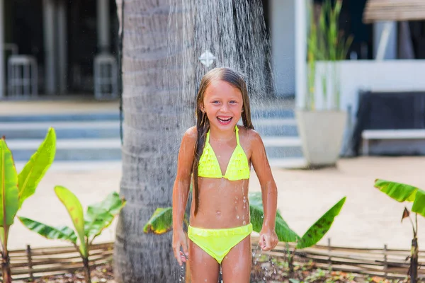 Hermosa niña feliz en un traje de baño amarillo bajo una ducha en la playa en un jardín tropical — Foto de Stock