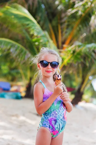 Gadis kecil yang lucu berkacamata hitam dengan es krim di pantai dengan pohon palem di laut. surga . — Stok Foto