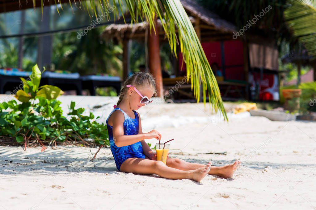 little cute girl on the beach in a bathing suit, sunglasses, sitting under a palm tree, drinking exotic cocktail
