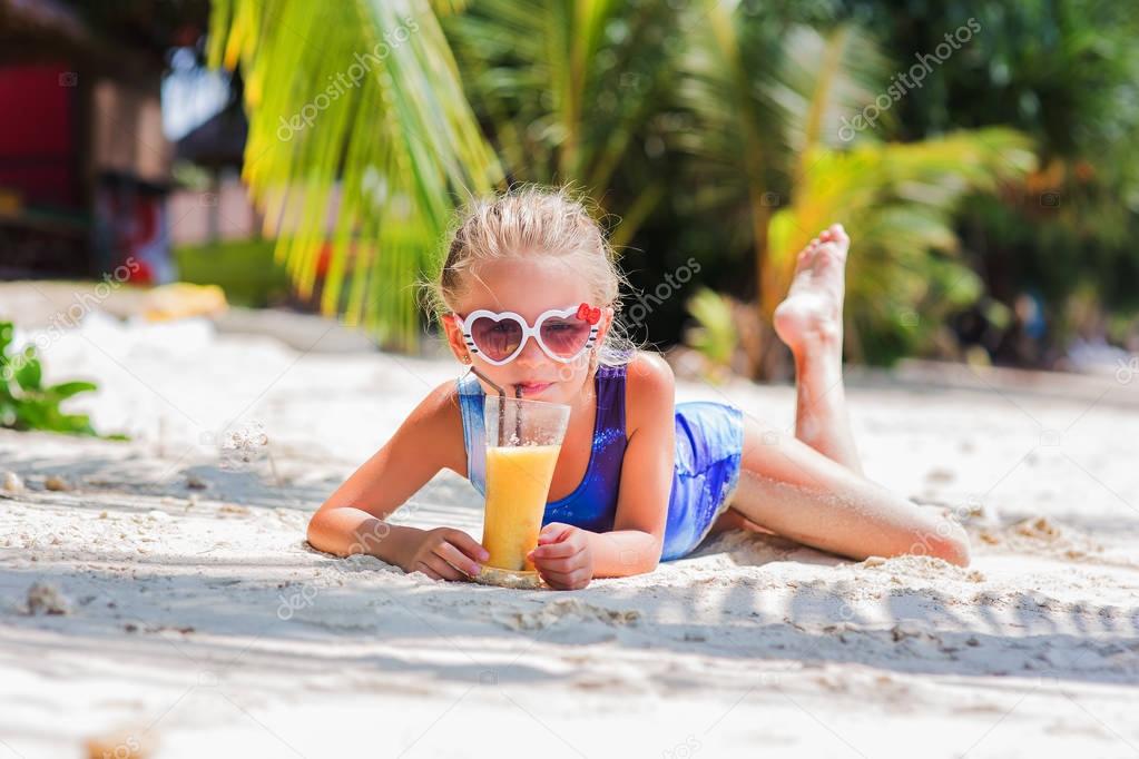 little cute girl on the sand at the beach in sun glasses with a glass of exotic cocktail juice