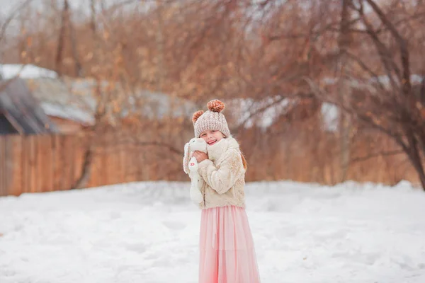 Miúda sorrindo bonito segurando coelho andando na neve no parque. A olhar para a câmara. Infância. Usando chapéu de malha, suéter e saia ao ar livre . — Fotografia de Stock