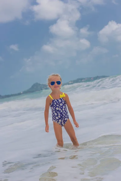 Маленькая девочка в купальнике и солнцезащитных очках на пляже на синем берегу моря в летние каникулы в дневное время — стоковое фото