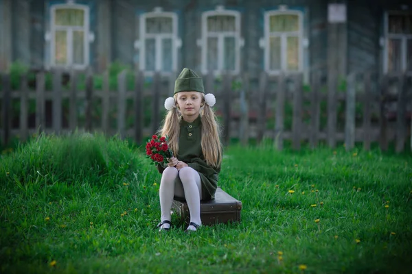 Nettes Mädchen in Militäruniform sitzt auf einem alten Koffer mit Blumen im Dorf — Stockfoto