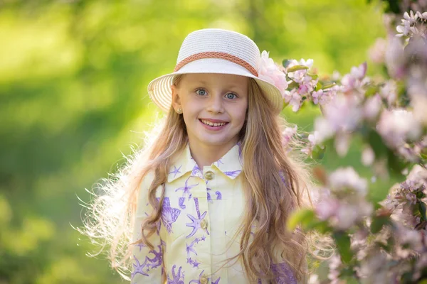 Schattig weinig gelukkig klein meisje met een hoed op een achtergrond van een bloeiende boom in een park — Stockfoto