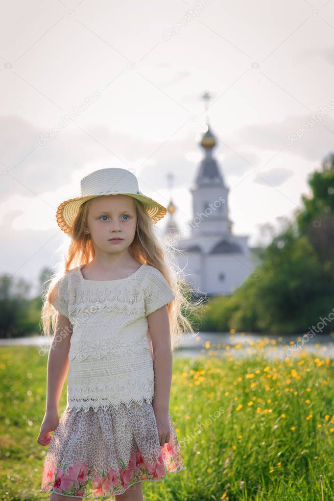 Portrait of a cute little girl in a hat on a summer day against the backdrop of a church in the village
