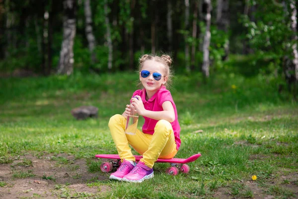 Une jolie petite fille en lunettes de soleil s'assoit sur une planche à roulettes dans une journée d'été dans le parc — Photo