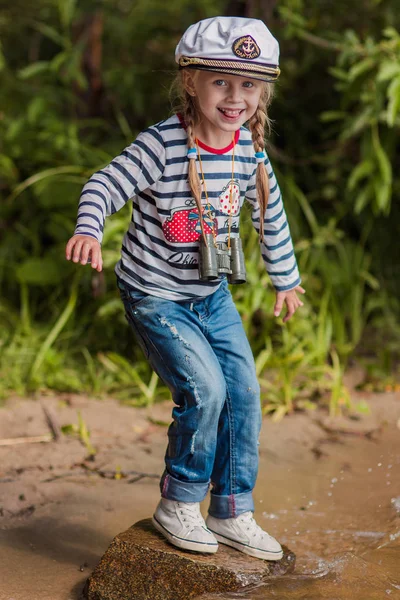 Ein kleines fröhliches Mädchen in Matrosenuniform und Seemütze steht am Ufer — Stockfoto