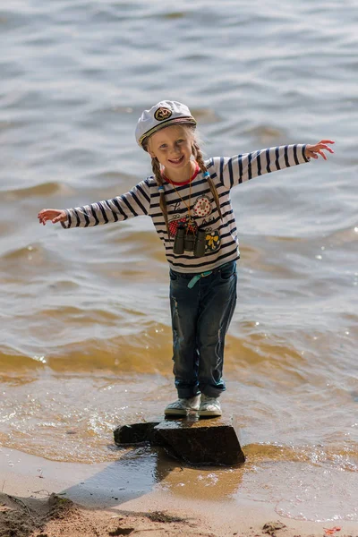 Una pequeña niña alegre en el uniforme de un marinero y una gorra de mar se encuentra en una gran roca en el río. El concepto de infancia. Concepto de descanso —  Fotos de Stock