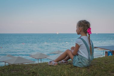 Küçük kız sahilde oturuyor ve denize bakarak