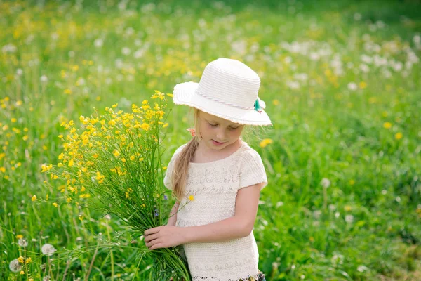 Little beautiful smiling blonde child girl 4-5 years in hat with fields in the field with yellow flowers, with a bouquet in hands — Stock Photo, Image