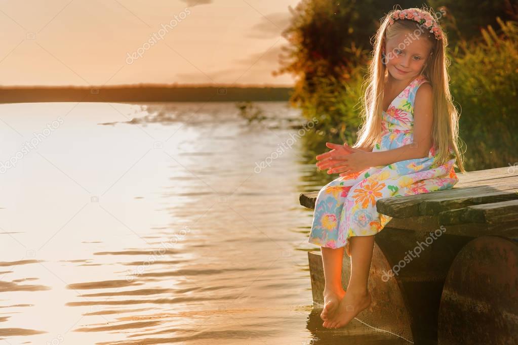Beautiful cute girl with long hair sitting on a bridge on the river bank in the summer evening at sunset