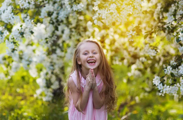 Een schattige mooie meisje met lang haar lacht naast een bloeiende boom in een park bij zonsondergang — Stockfoto