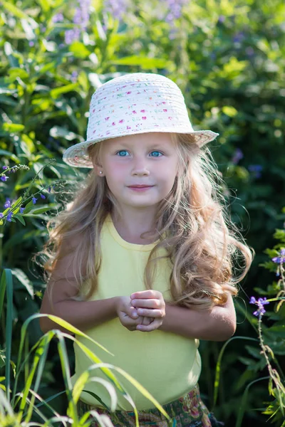 Souriant mignon bébé fille 4-5 ans tenant fleur portant un grand chapeau dans la prairie. Je regarde la caméra. Enfance. Heure d "été . — Photo