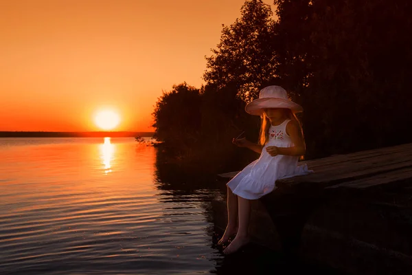 Little girl with a white dress and a white hat sits on a bridge on the lake at sunset in the summer. Holidays. Recreation — Stock Photo, Image