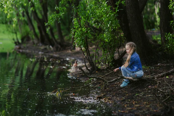 A menina está sentada junto à lagoa com um pau nas mãos. Férias. Recreação — Fotografia de Stock