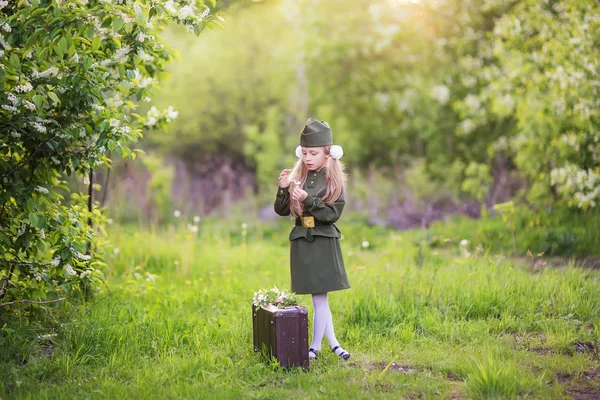 Den vítězství. Dívka v šatech vojáka stojí vedle kufr v trávě pod stromem — Stock fotografie