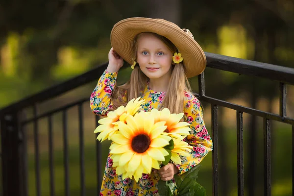 Ritratto ravvicinato di Bella bambina bionda in abito giallo godendo sera. Ragazza sorridente felice in un cappello con girasole di fiori . — Foto Stock