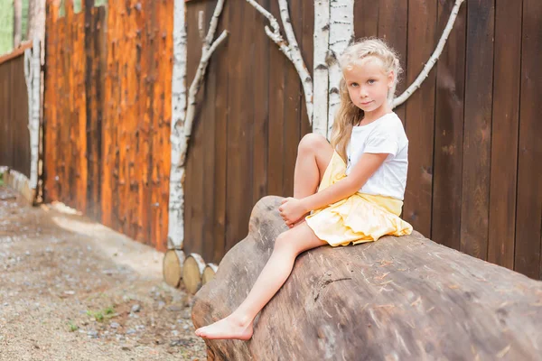 Χαριτωμένο μικρό κορίτσι που κάθεται σε ένα μεγάλο κούτσουρο μέρα του καλοκαιριού — Φωτογραφία Αρχείου