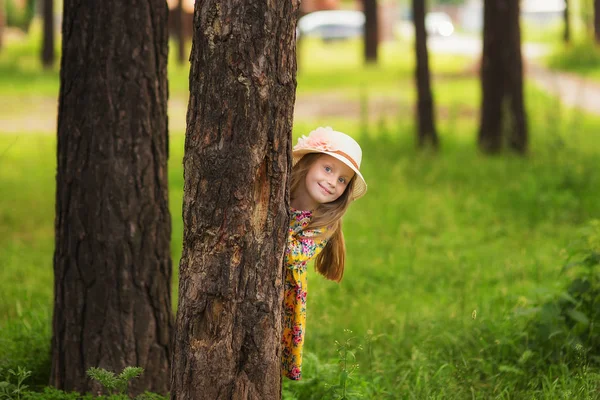 Doce menina bonita em um chapéu brincando espreitando atrás de uma árvore. Ri menina . — Fotografia de Stock